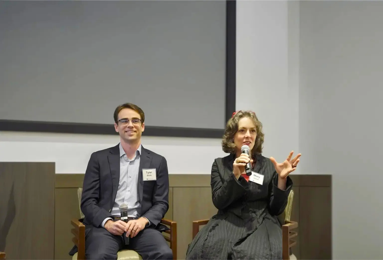 Two people sit on chairs at a panel discussion; one holds a microphone and gestures while speaking, while the other sits with hands folded, both wearing name tags. The discussion features insights from San Jose IRS whistleblower lawyers as they address questions about tax law and whistleblower protections.