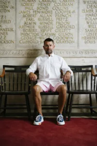 Man in a white shirt and pink shorts sits on a black bench against a marble wall inscribed with names in gold lettering.