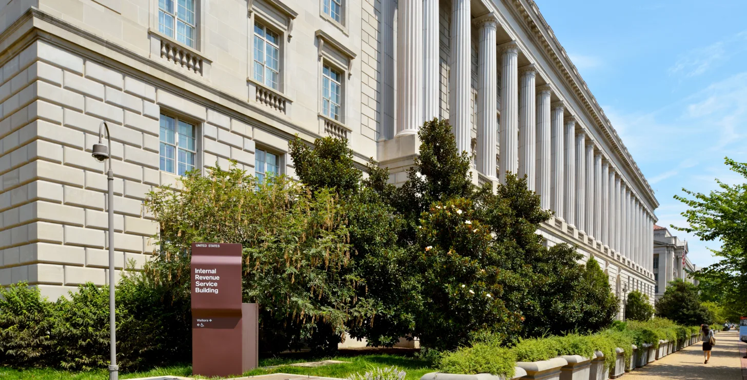 A large neoclassical building with tall columns and a sign reading “Internal Revenue Service Building” along a landscaped sidewalk on a sunny day.