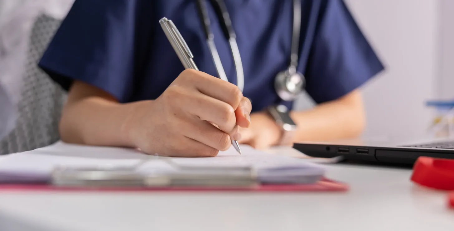 Person in blue medical scrubs and a stethoscope writes on paper at a desk near a laptop, consulting with a Silicon Valley FCA whistleblower lawyer.